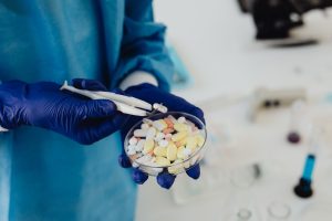Gloved hands examine assorted tablets and capsules in a petri dish
