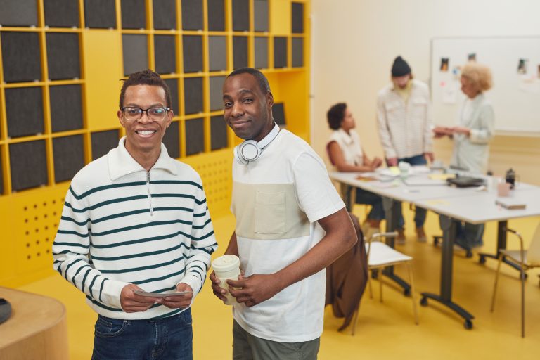 Two coworkers stand in a bright startup workspace while teammates collaborate behind them